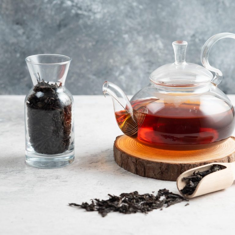 Thé noir infusé dans une théière en verre avec des feuilles sèches à côté.