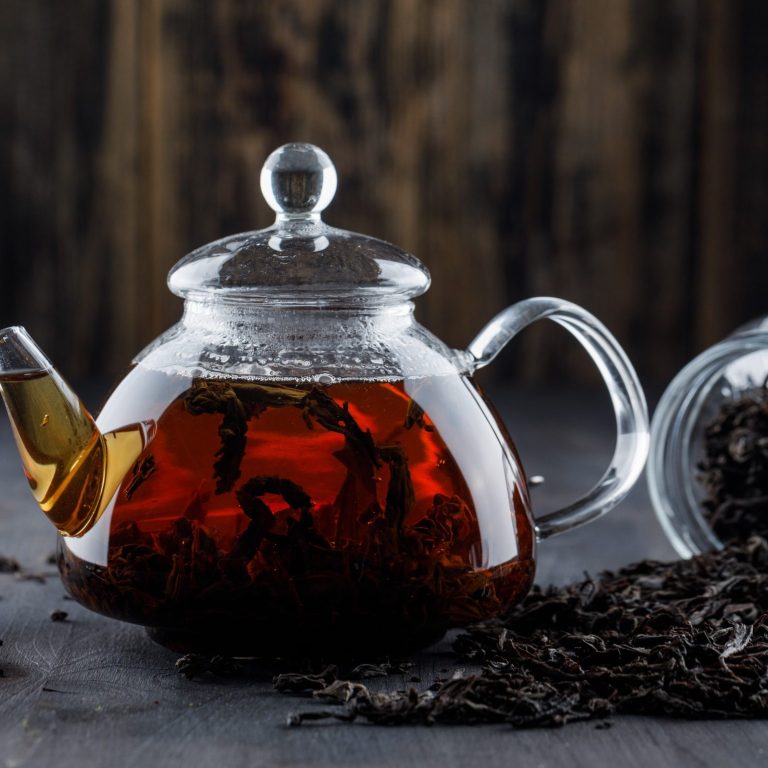 Théière en verre remplie de thé noir, avec des feuilles de thé à côté.